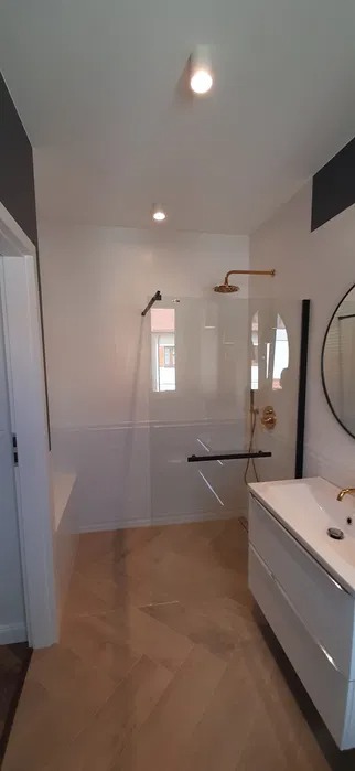 Nowoczesna łazienka z kabiną prysznicową typu walk-in, złotą armaturą, białymi płytkami do połowy ściany i parkietem ułożonym w jodełkę.