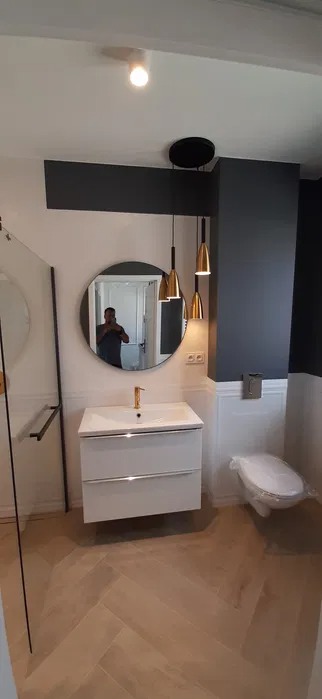 Wykończona łazienka z parkietem ułożonym w jodełkę, białą szafką pod umywalką z złotą baterią, okrągłym lustrem, wiszącymi złotymi lampami i toaletą owiniętą folią ochronną.