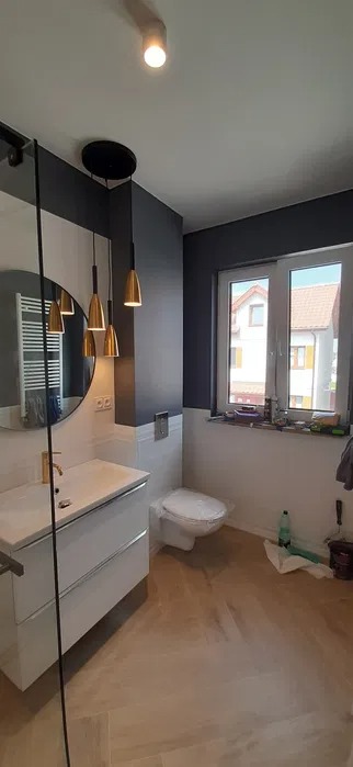 Nowoczesna łazienka z elementami złota: lampy wiszące, okrągłe lustro, biała umywalka z szafką, szare ściany w górnej części, białe płytki do połowy wysokości, beżowa podłoga imitująca drewno...