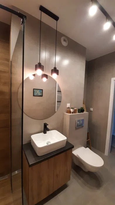 Nowoczesna łazienka z umywalką nablatową na drewnianej szafce, okrągłym lustrem z podświetleniem LED, wiszącą toaletą i minimalistycznym oświetleniem punktowym.