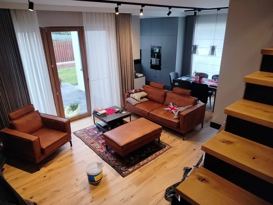 Jasne drewniane schody prowadzą do salonu z brązową skórzaną sofą, fotelem i pufem, widoczne ślady po remoncie: wiadro po farbie na dywanie, w tle szara zabudowa kuchenna i stół jadalny.