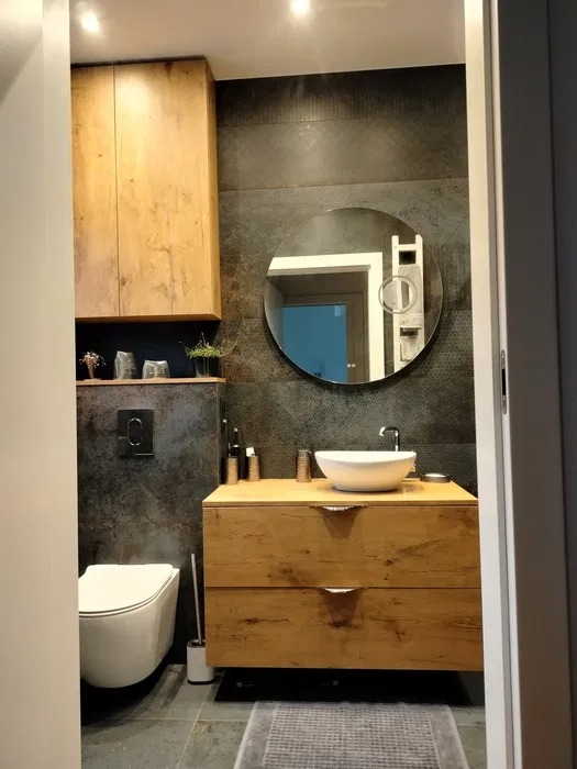 Nowoczesna łazienka z drewnianą szafką pod umywalką, okrągłym lustrem i ciemnymi płytkami na ścianie, widok zza framugi drzwi.