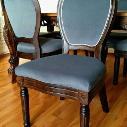 A&K KOCIEMBA SPÓŁKA JAWNA - Krzesło z ciemnego drewna z szarym tapicerowanym siedziskiem i oparciem, ozdobne rzeźbienia na nogach i ramie, biała lamówka wokół oparcia, parkiet na podłodze.