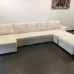 Narożnik modułowy z białego sztruksu z pufami, na ciemnej podłodze z płytek, plakat na ścianie w tle.