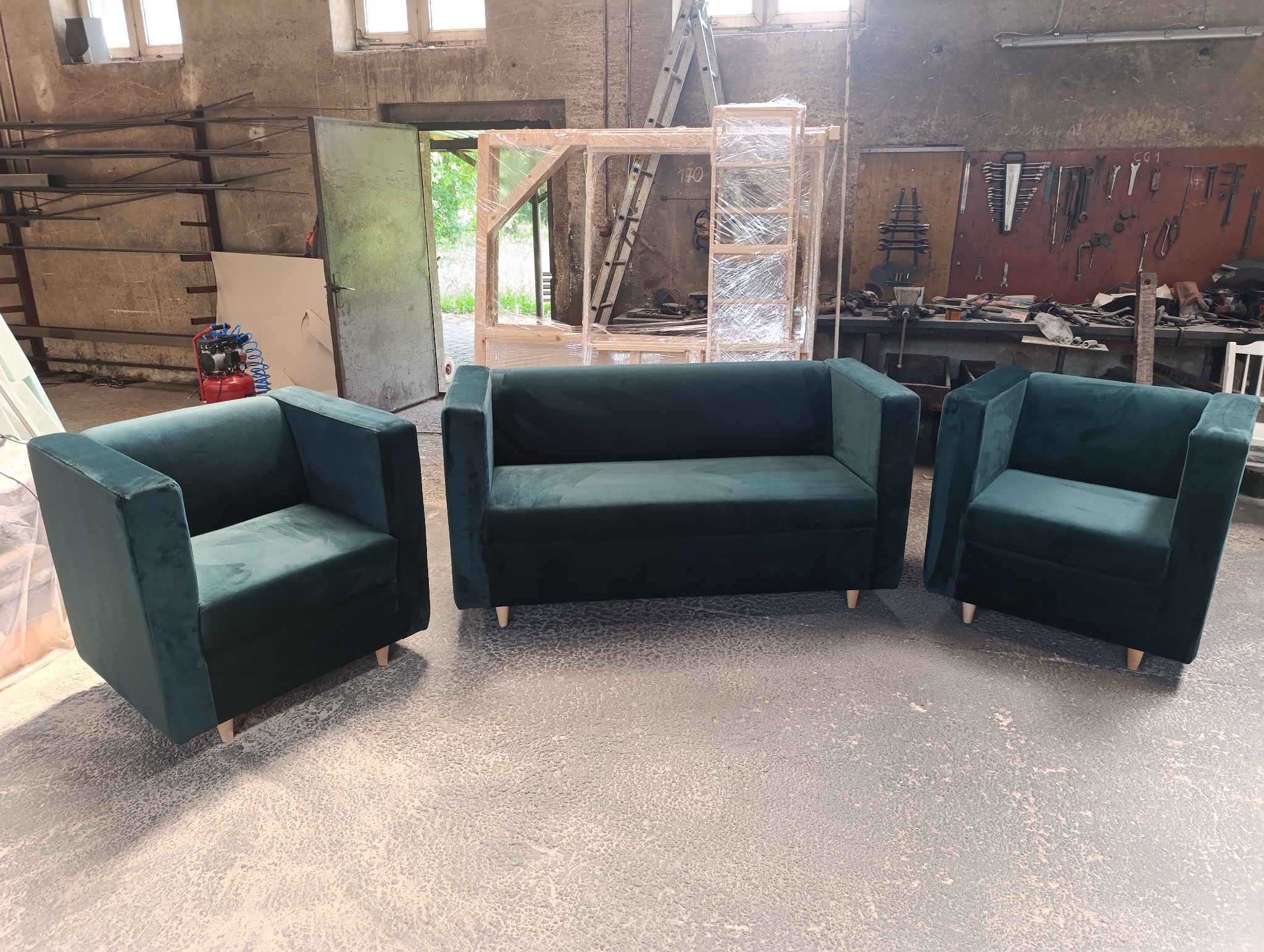 Komplet wypoczynkowy: sofa dwuosobowa i dwa fotele w kolorze butelkowej zieleni, na tle warsztatu z narzędziami i ramą meblową w folii.