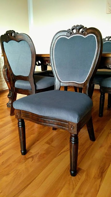 Krzesło z ciemnego drewna z szarym tapicerowanym siedziskiem i oparciem, ozdobne rzeźbienia na nogach i ramie, biała lamówka wokół oparcia, parkiet na podłodze.