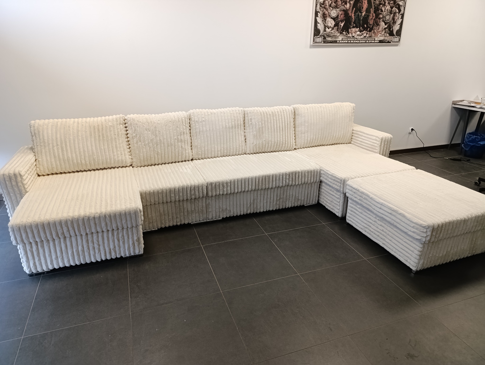 Narożnik modułowy z białego sztruksu z pufami, na ciemnej podłodze z płytek, plakat na ścianie w tle.