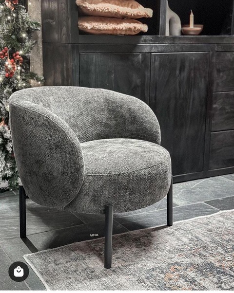 Szary fotel tapicerowany o zaokrąglonym kształcie na czarnych, metalowych nóżkach, ustawiony na wzorzystym dywanie w pomieszczeniu z ciemnymi meblami i choinką w tle.