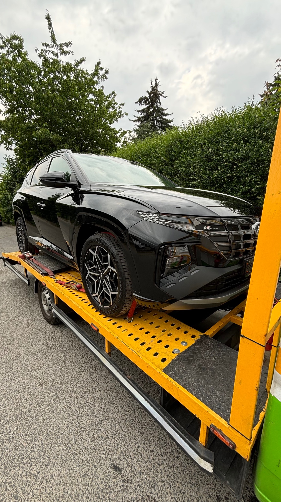 Czarny Hyundai Tucson N Line zabezpieczony czerwonymi pasami na żółtej lawecie, widok z przodu na tle zieleni i pochmurnego nieba.