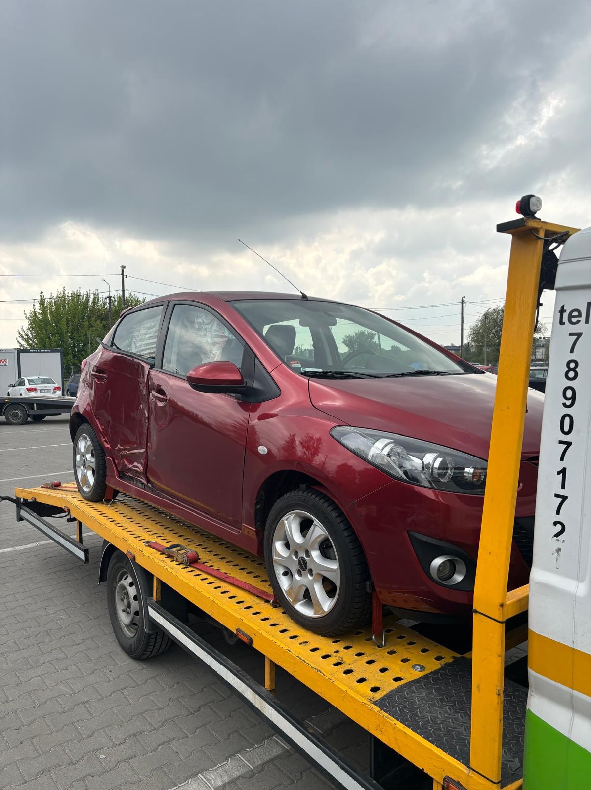Czerwony uszkodzony samochód Mazda na żółtej platformie lawety, widoczne wgniecenia na drzwiach i błotniku, pochmurne niebo w tle.