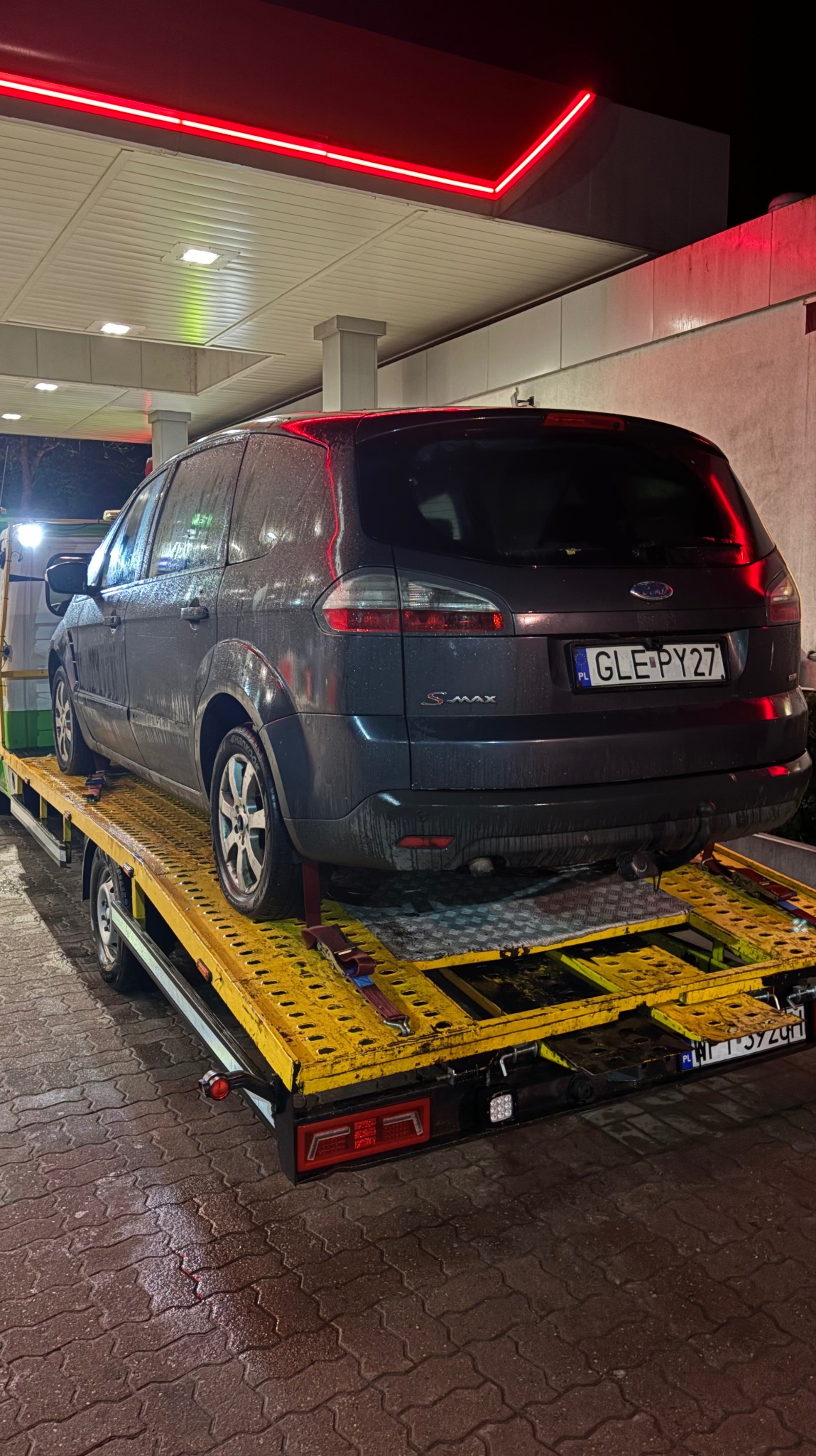 Szary samochód Ford S-Max z polską tablicą rejestracyjną GLE PY27 umieszczony na żółtej platformie lawety, pod zadaszeniem stacji benzynowej z czerwonym neonowym oświetleniem.