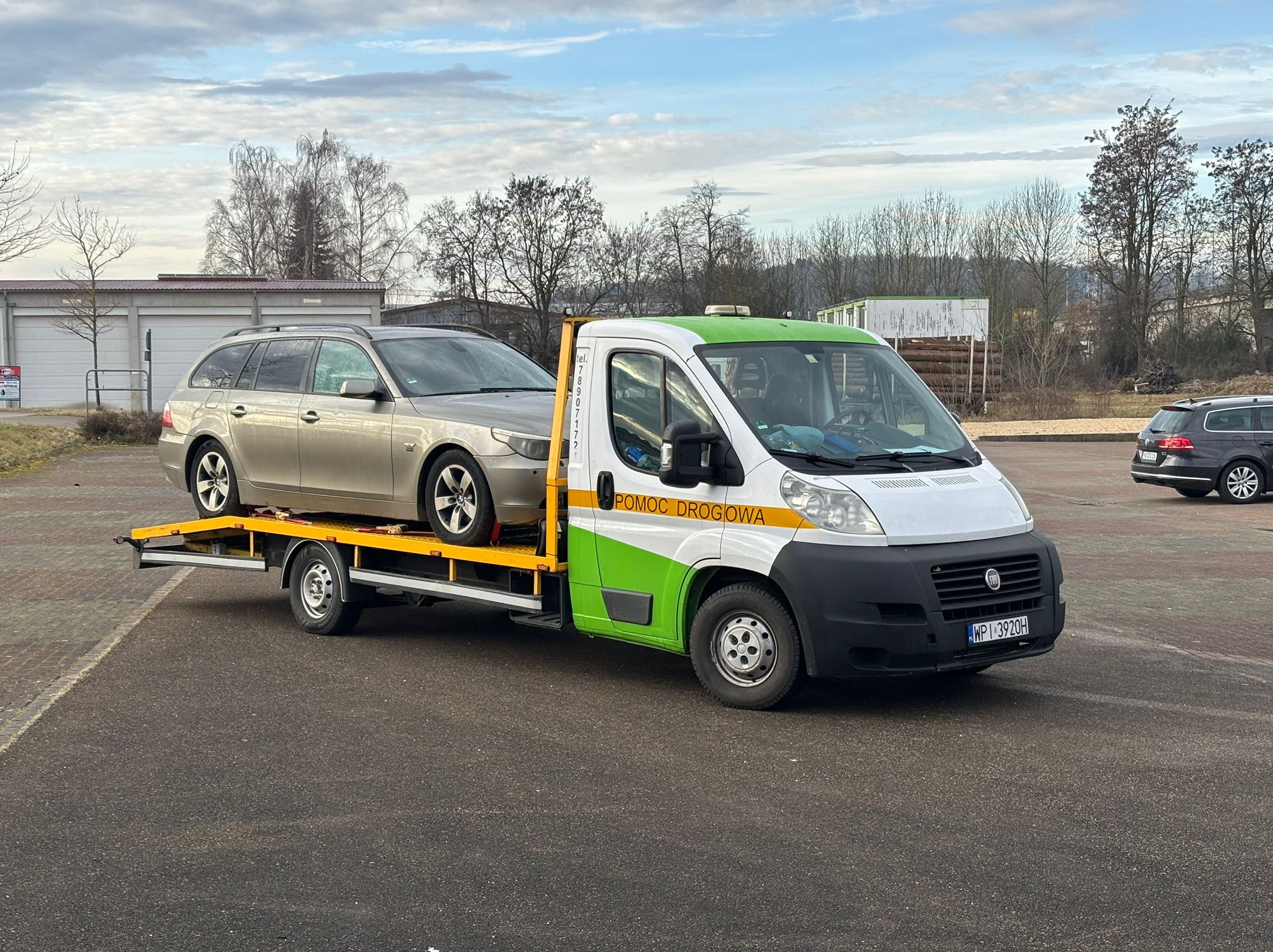 Samochód osobowy w kolorze szampańskim na żółtej platformie lawety pomocy drogowej z zielonymi akcentami na tle parkingu i drzew bez liści.