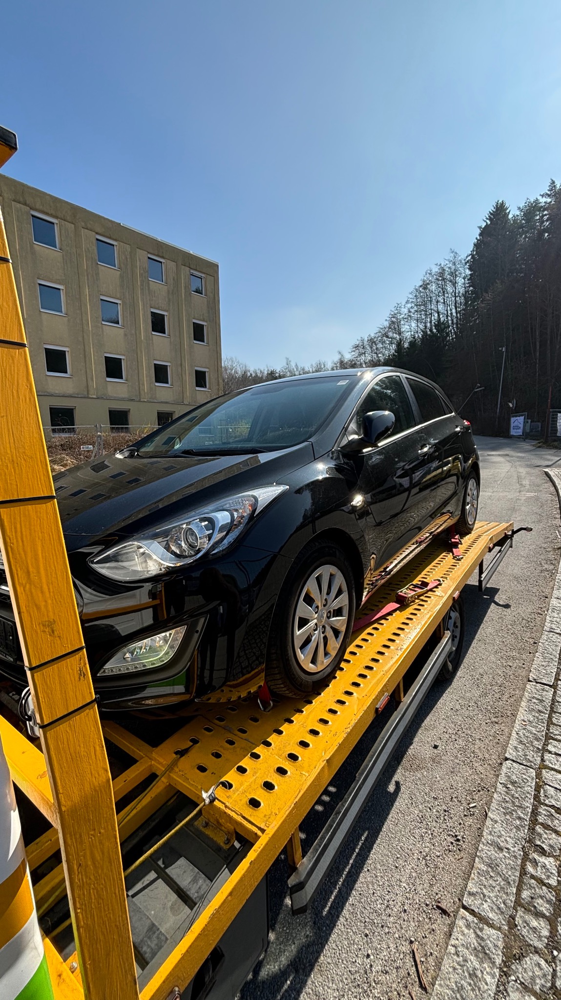 Czarny samochód osobowy zabezpieczony pasami transportowymi na żółtej platformie lawety, w tle budynek i drzewa w słoneczny dzień.