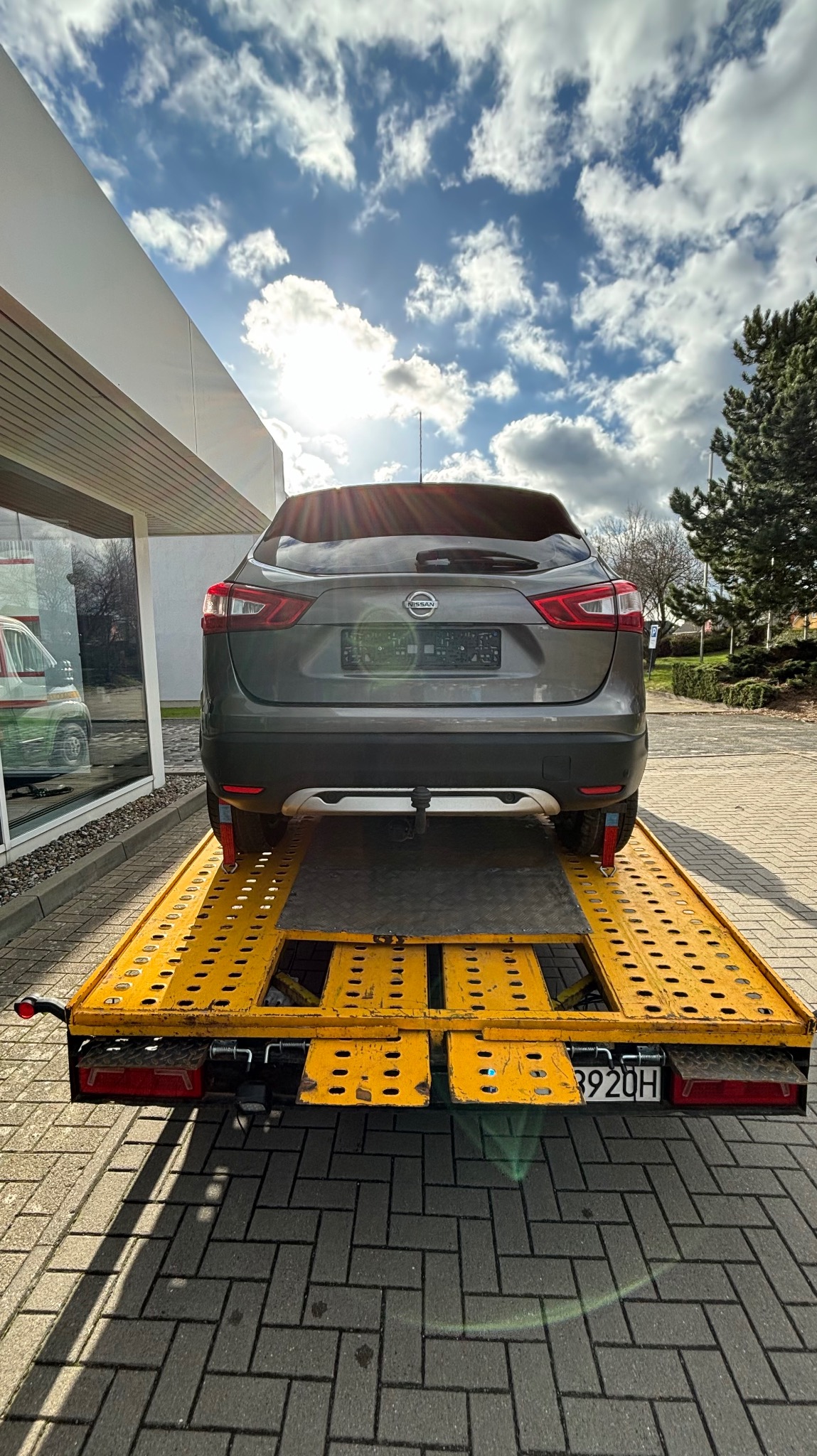 Szary Nissan Qashqai zabezpieczony czerwonymi pasami na żółtej, ażurowej lawecie, z widoczną tablicą rejestracyjną, na tle kostki brukowej i budynku z refleksem innego pojazdu w szybie