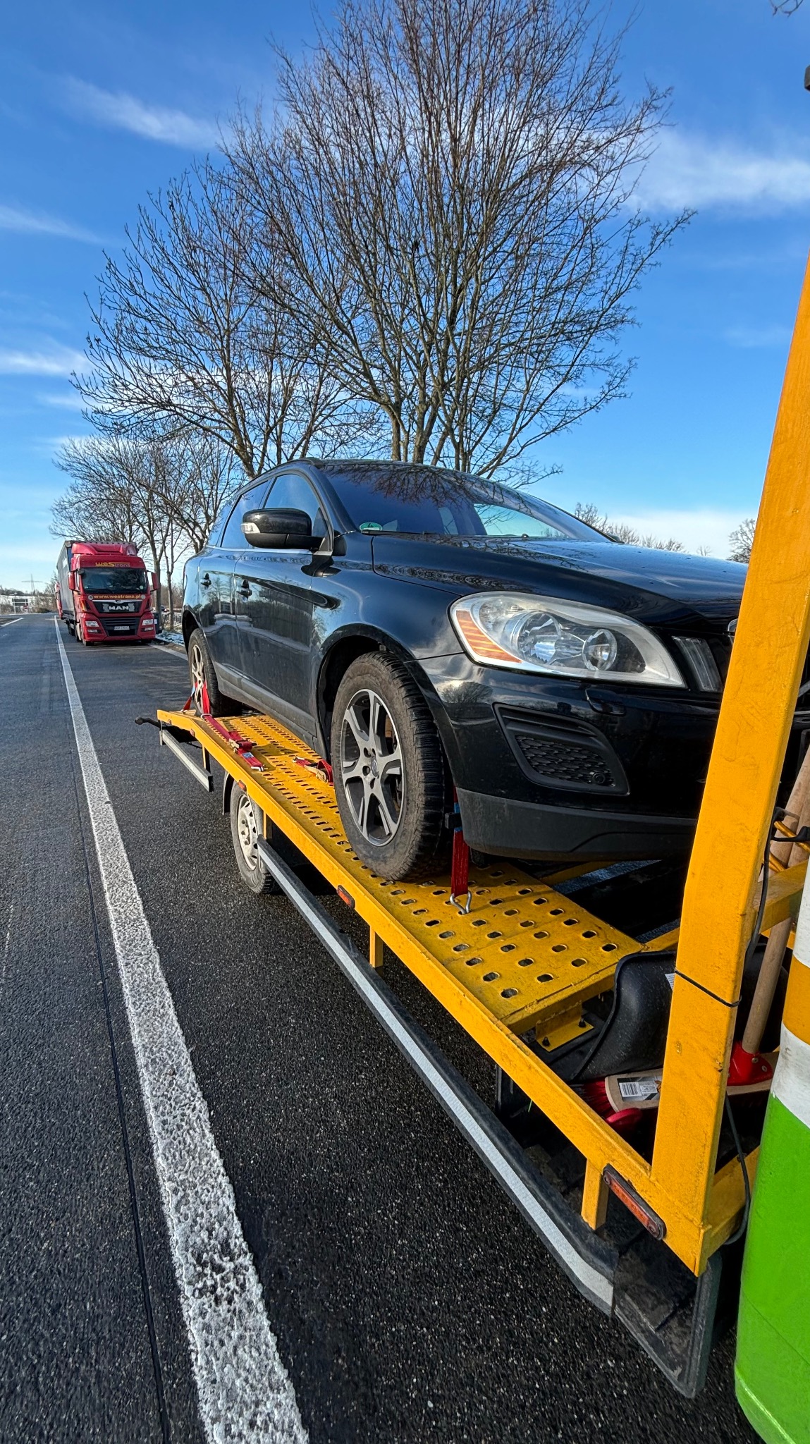Czarny samochód osobowy Volvo na żółtej platformie lawety, zabezpieczony czerwonymi pasami transportowymi, widok z boku na autostradzie w słoneczny dzień.