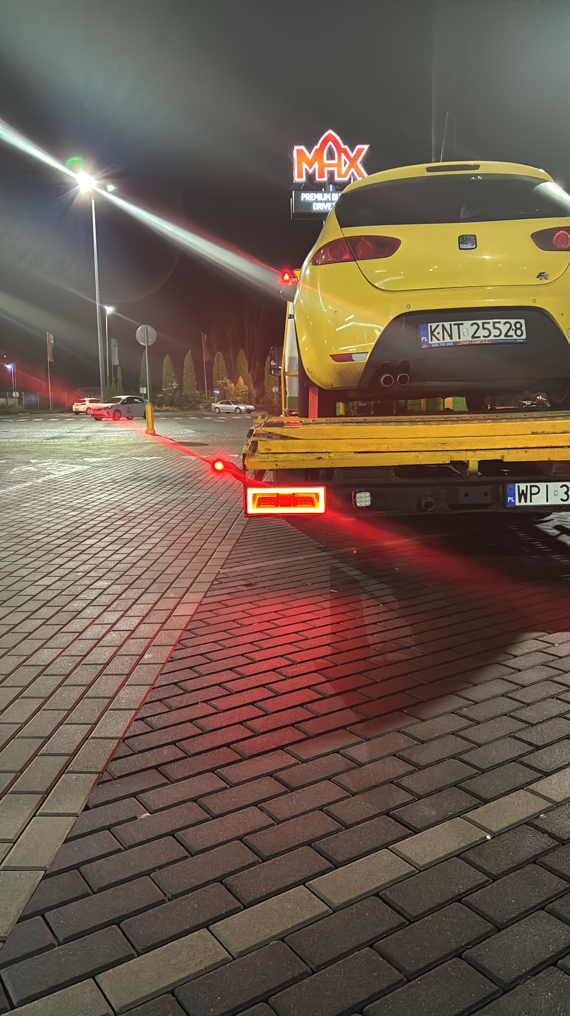 Żółty samochód marki Seat na żółtej lawecie, widok z tyłu, nocą, na parkingu przed sklepem MAX Premium Drive, z włączonymi światłami lawety.