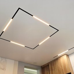 REMFIX Sufity Napinane - Nowoczesny sufit z podświetleniem LED w kształcie kwadratu i linii, czarne obramowanie, ciepłe światło, minimalistyczny design, fragment kuchni w tle.