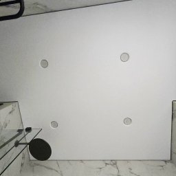 REMFIX Sufity Napinane - Biały sufit napinany z czterema punktami świetlnymi w łazience wykończonej marmurem, widok z dołu, fragment kabiny prysznicowej i lustra w złotej ramie.