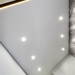 REMFIX Sufity Napinane - Biały sufit z oświetleniem LED i punktowymi lampami, widok pod kątem. Dekoracyjna tapeta z motywem liści w rogu pomieszczenia. Nowoczesne wykończenie wnętrza.