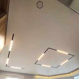 REMFIX Sufity Napinane - Nowoczesny sufit z geometrycznymi oprawami LED w kształcie kwadratu i linii, oświetlony pokój z dużymi oknami. Minimalistyczny design, jasne wnętrze.
