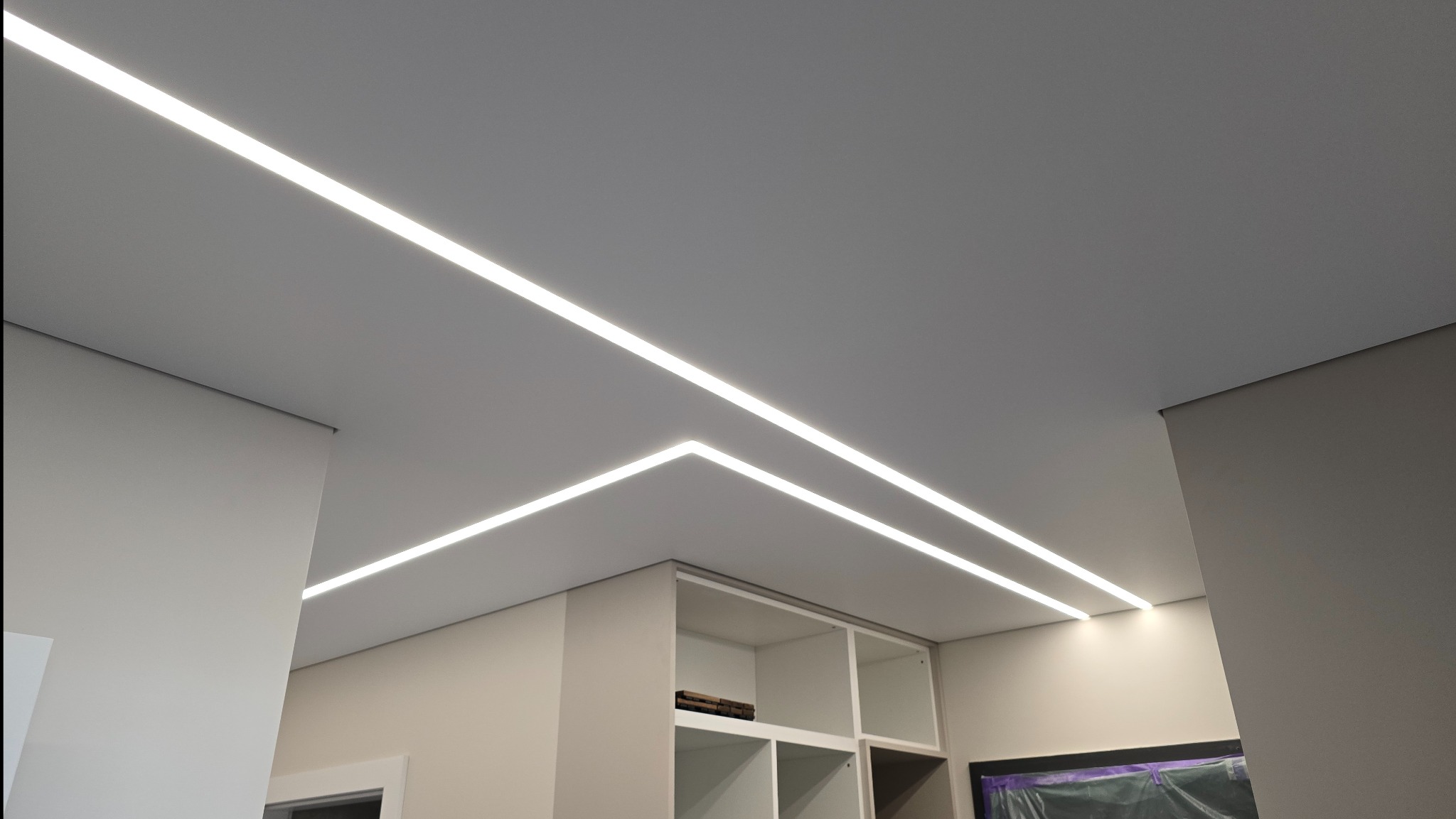Minimalistyczny, szary sufit z wbudowanym oświetleniem LED w postaci długich, prostych linii, tworzących geometryczny wzór i równomiernie oświetlających wnętrze. Widoczny fragment zabudowy meblowej...