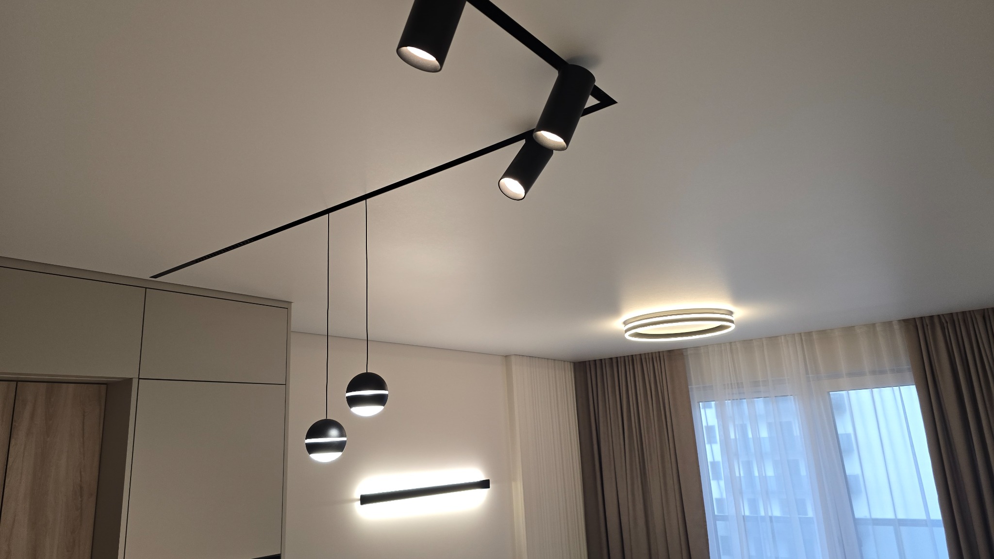 Biały, gładki sufit z zamontowanym systemem szynoprzewodów z reflektorami, okrągłą lampą sufitową i wiszącymi lampami kulami. Nowoczesne oświetlenie w salonie.