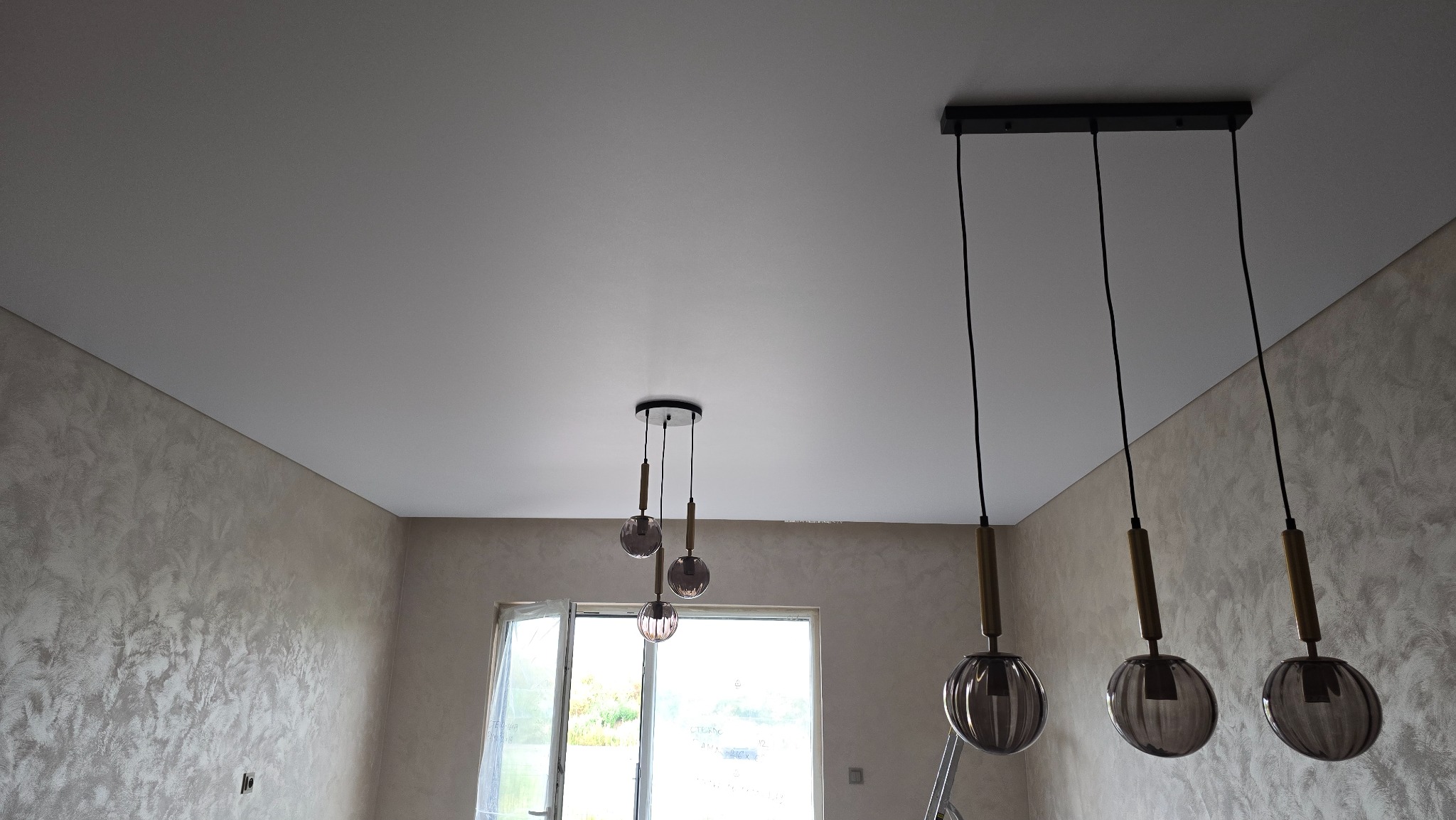 Gładki, biały sufit po montażu, z nowoczesnymi lampami wiszącymi w pomieszczeniu z dekoracyjną ścianą i oknem.