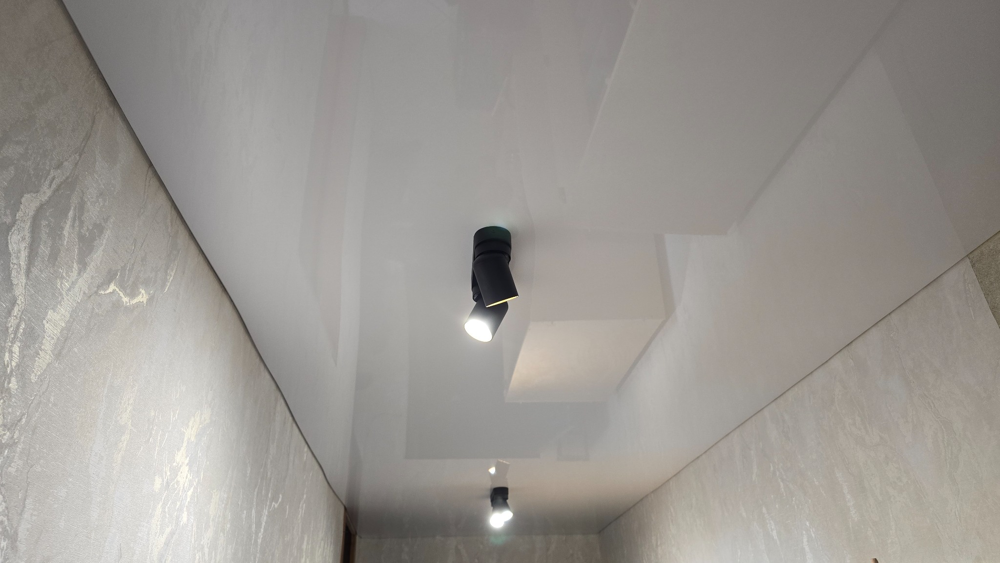 Biały, błyszczący sufit napinany z czarnymi reflektorami punktowymi, zamontowany w nowoczesnym wnętrzu z jasnymi ścianami o delikatnej strukturze.