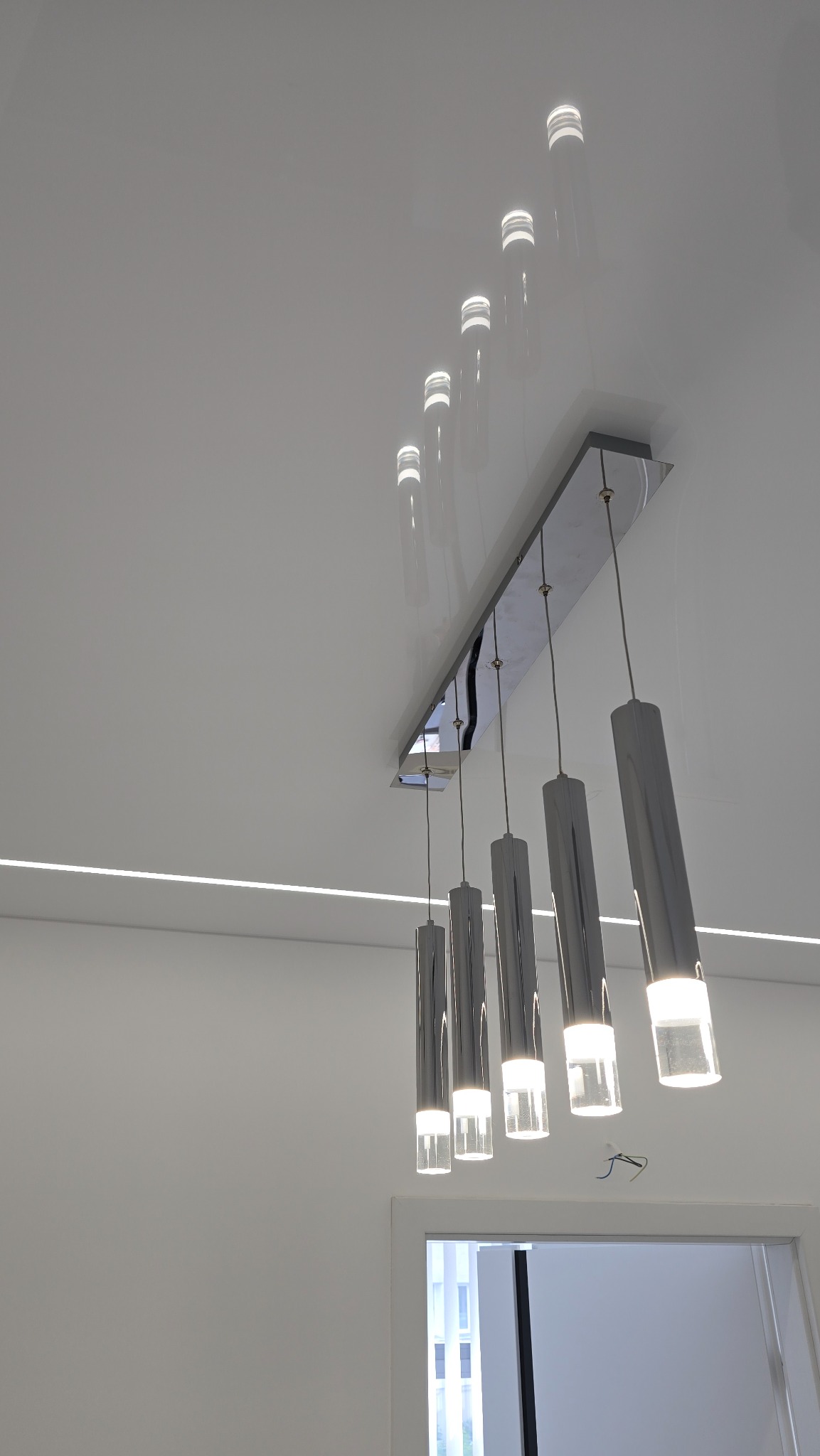 Biały, połyskujący sufit z listwą LED i nowoczesną lampą wiszącą z pięcioma chromowanymi tubami. Widok z dołu, minimalistyczny design.