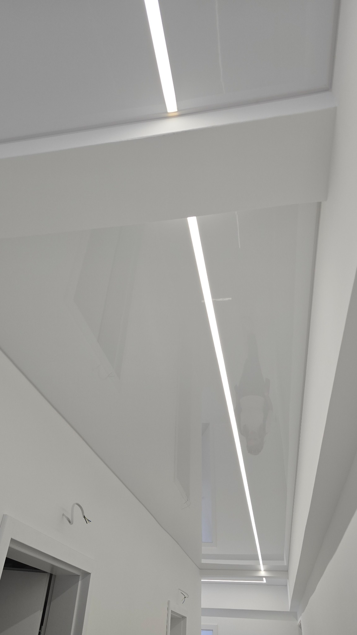 Biały, błyszczący sufit napinany z liniowym oświetleniem LED, widok z dołu, odbicie sylwetki osoby na suficie, minimalistyczny design.