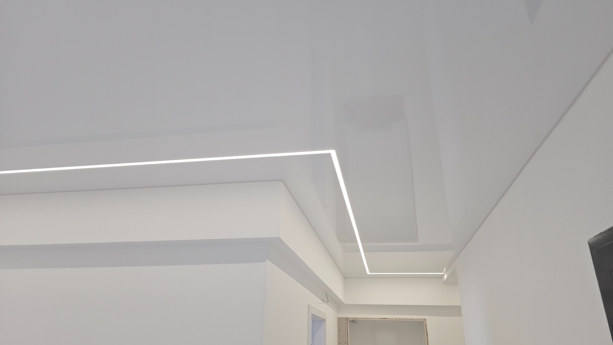 Biały, błyszczący strop z liniowym oświetleniem LED wzdłuż krawędzi, tworząc nowoczesny design. Minimalistyczne, jasne wnętrze z widocznymi elementami architektonicznymi.