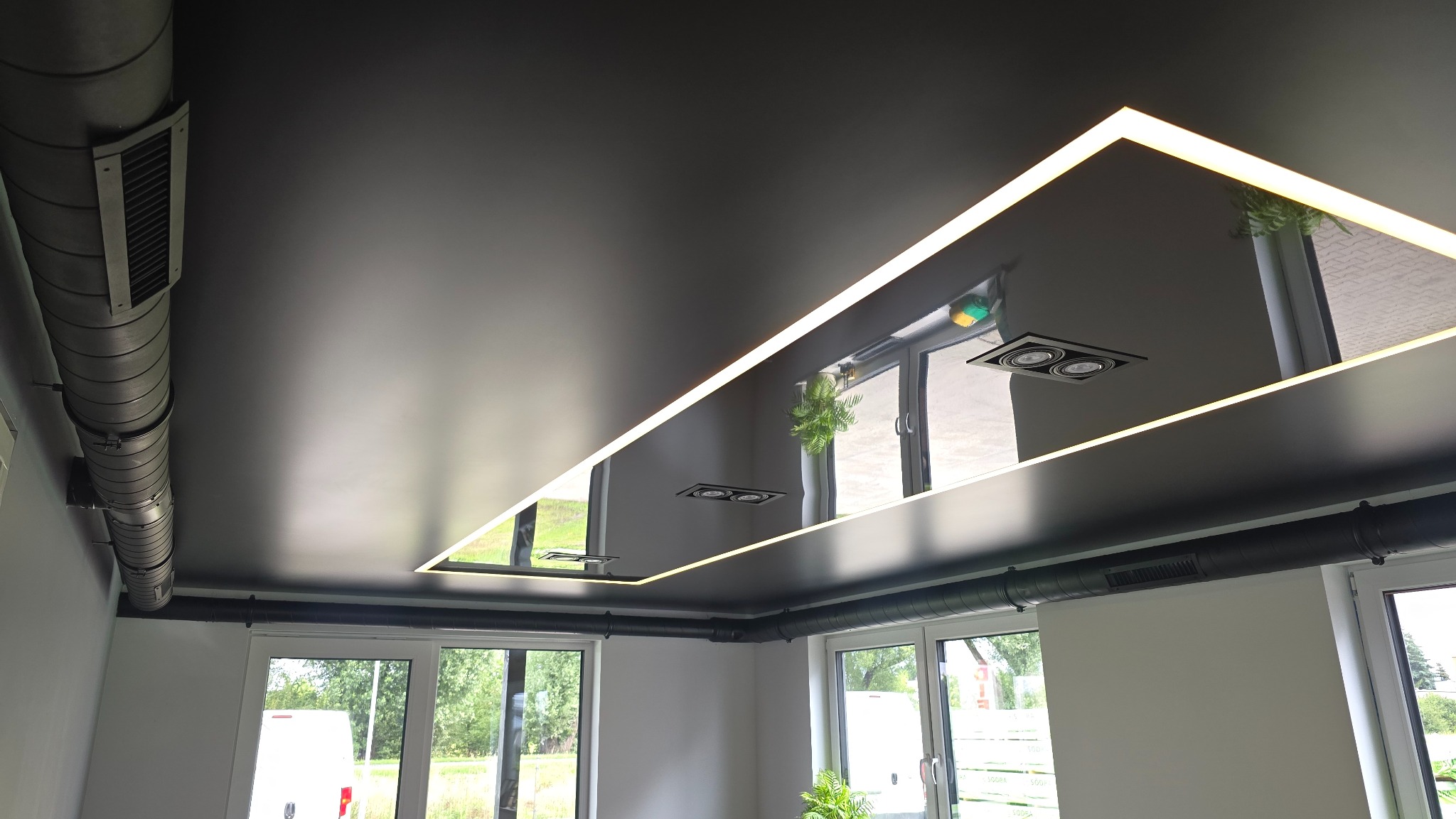 Błyszczący, czarny sufit z prostokątnym oświetleniem LED odbijającym otoczenie. Widoczne elementy wentylacji i okna.