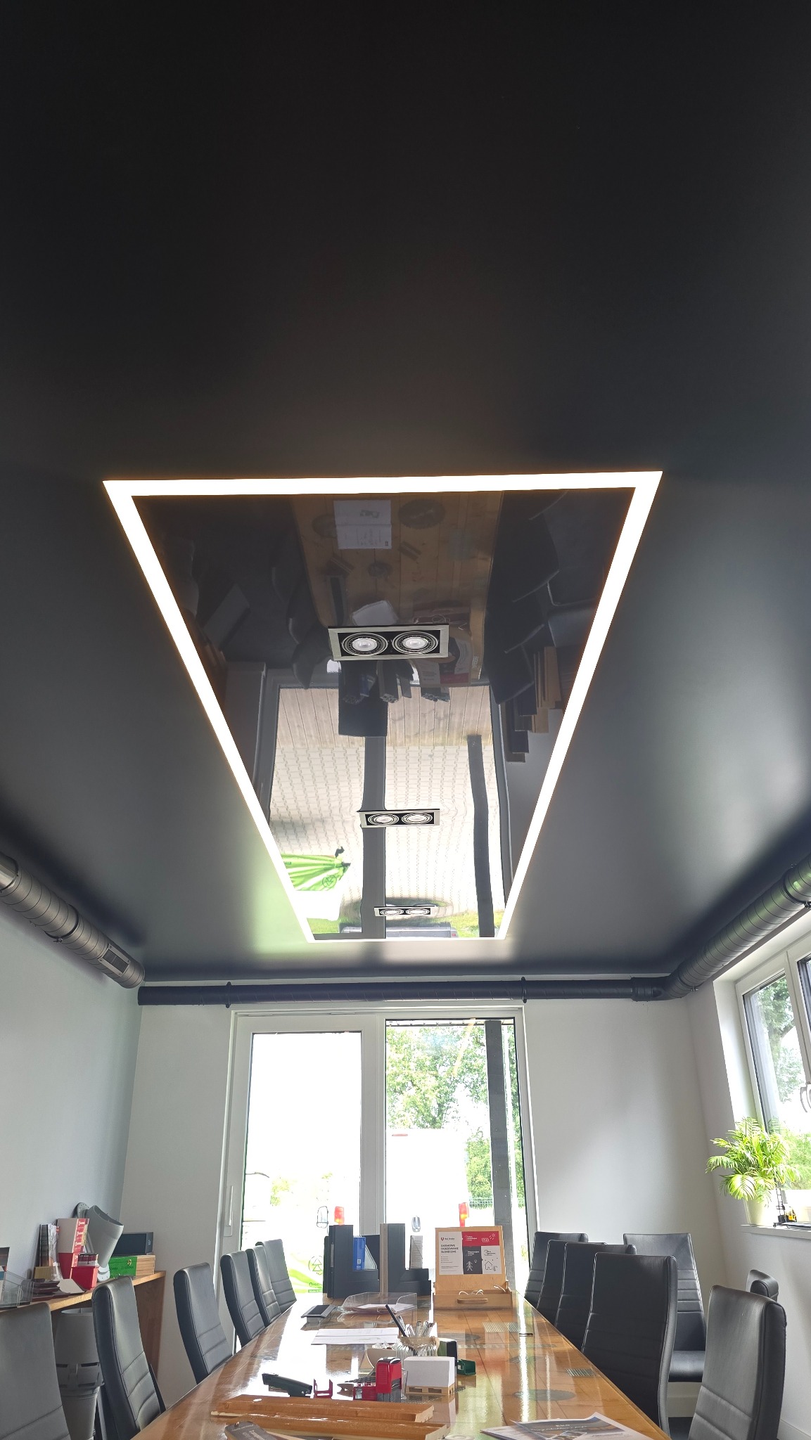 Nowoczesny, czarny sufit z trójkątnym oświetleniem LED odbijającym wnętrze biura. Widok na stół konferencyjny i okna.