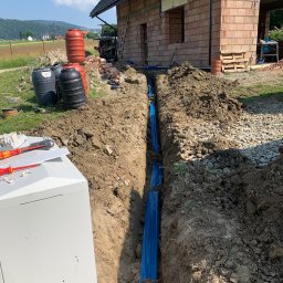 EL-DAW - Wykop z niebieską folią zabezpieczającą rury na tle budowy domu w stanie surowym otwartym, widoczne narzędzia elektryczne i zbiorniki na wodę.