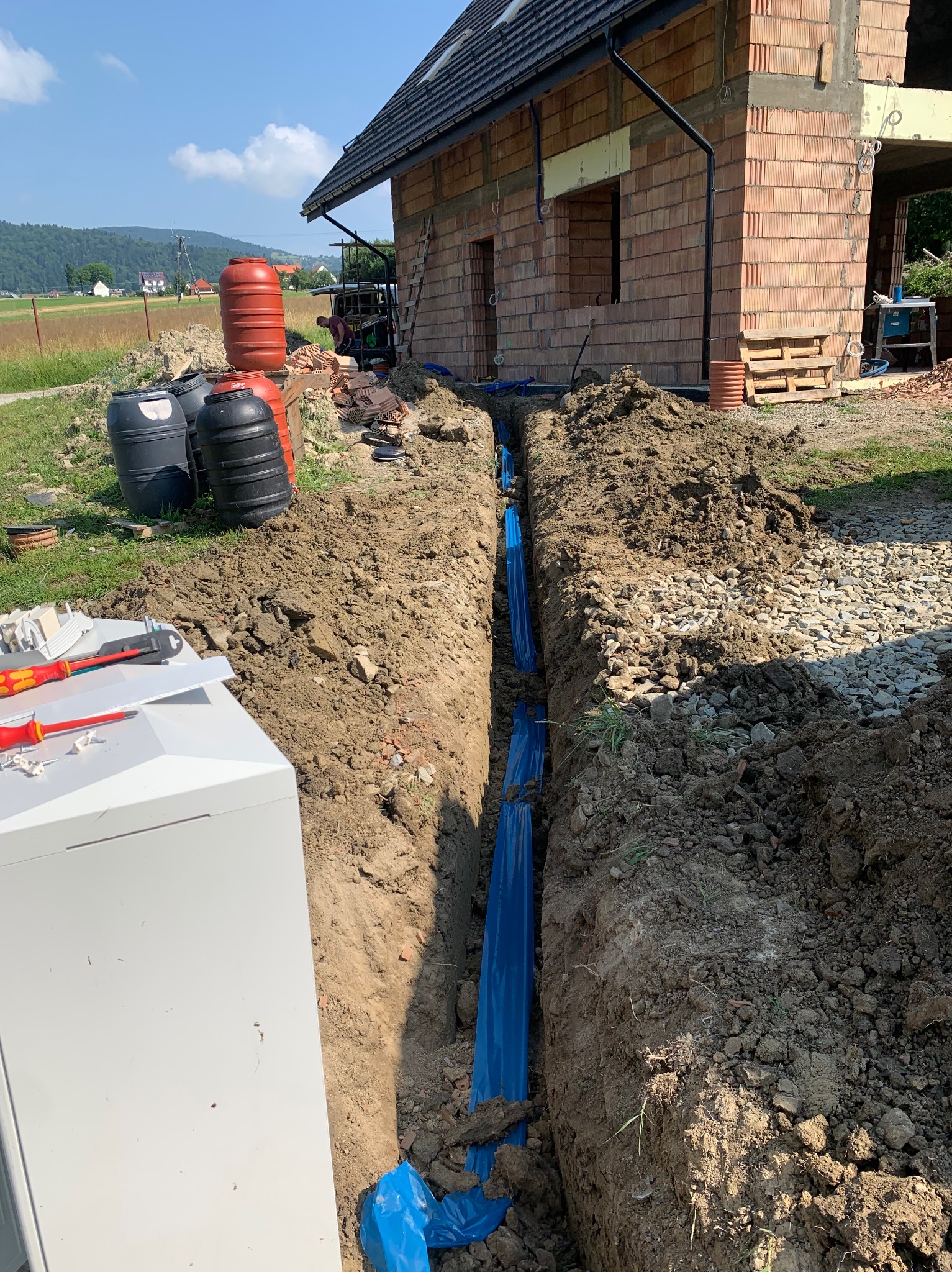 Wykop z niebieską folią zabezpieczającą rury na tle budowy domu w stanie surowym otwartym, widoczne narzędzia elektryczne i zbiorniki na wodę.
