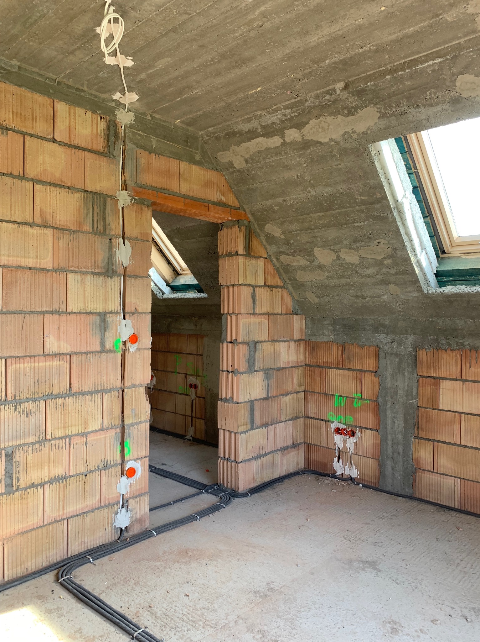 Wnętrze w trakcie budowy z widocznymi przewodami elektrycznymi w peszlach, prowadzącymi wzdłuż ściany z czerwonej cegły ceramicznej, z oznaczonymi punktami instalacyjnymi i surowym betonowym stropem.