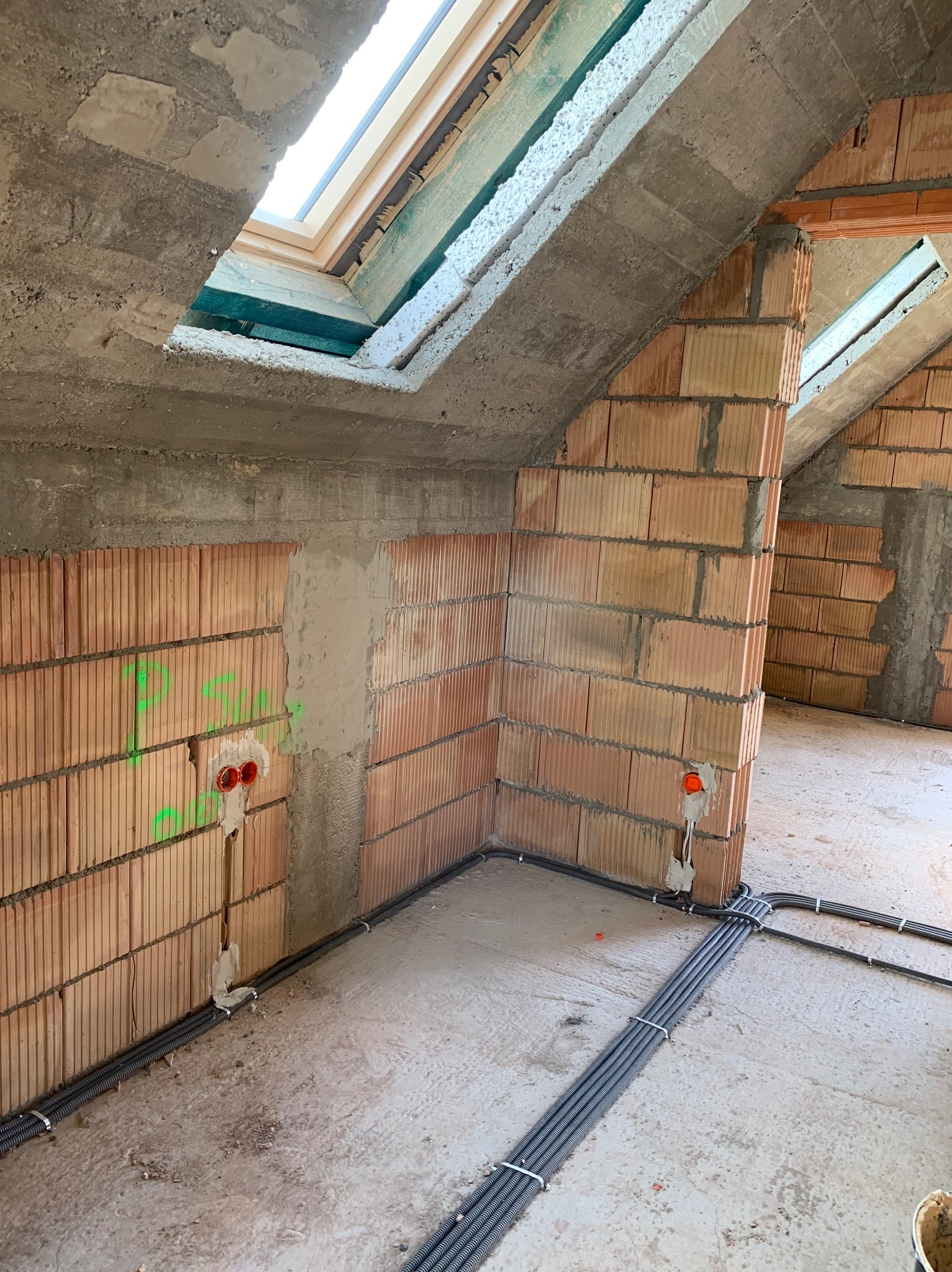 Surowy stan poddasza z widocznymi przewodami elektrycznymi w peszlach, zamontowanymi na ścianach z czerwonej cegły i podłodze betonowej, z oknem dachowym w tle.