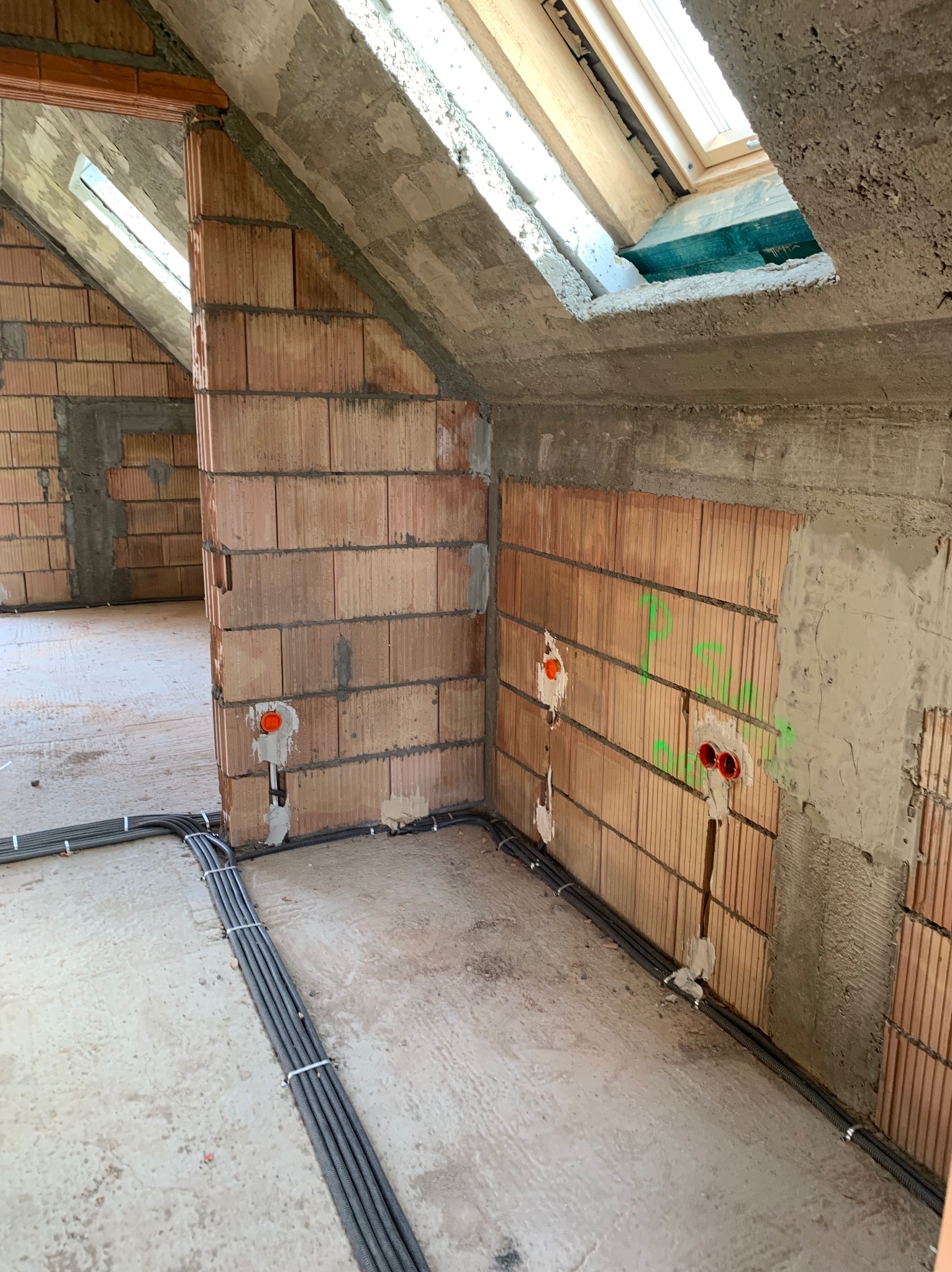 Surowy stan poddasza z widocznymi przewodami elektrycznymi w peszlach, wyprowadzonymi puszkami instalacyjnymi w ścianie z cegły dziurawki oraz oknem dachowym w betonowej ramie.
