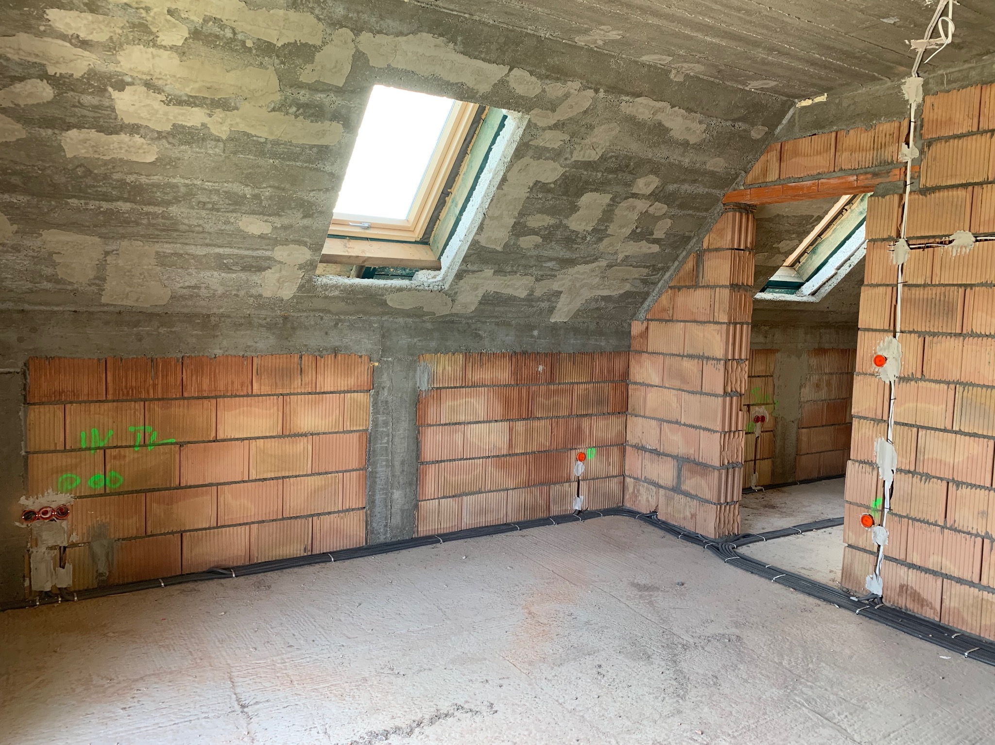 Surowy stan poddasza z oknami dachowymi, widoczne instalacje elektryczne w peszlach, ściany z czerwonej cegły z zaznaczonymi punktami elektrycznymi, surowy betonowy strop z widocznymi łatami.