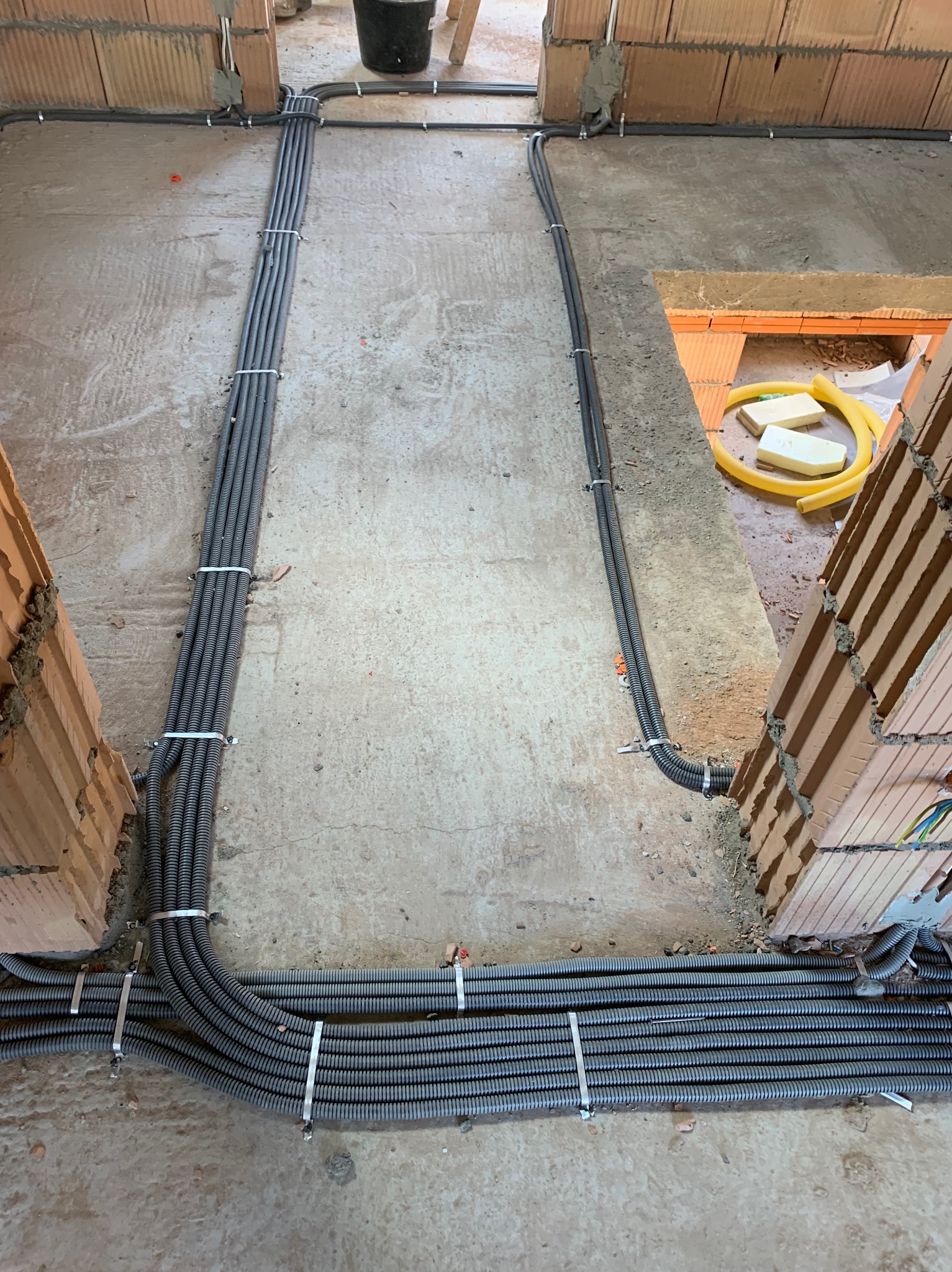 Szare rury instalacyjne ułożone w wiązki i przymocowane do surowej podłogi w budynku w Jurkowie, widoczne ceglane ściany w tle.