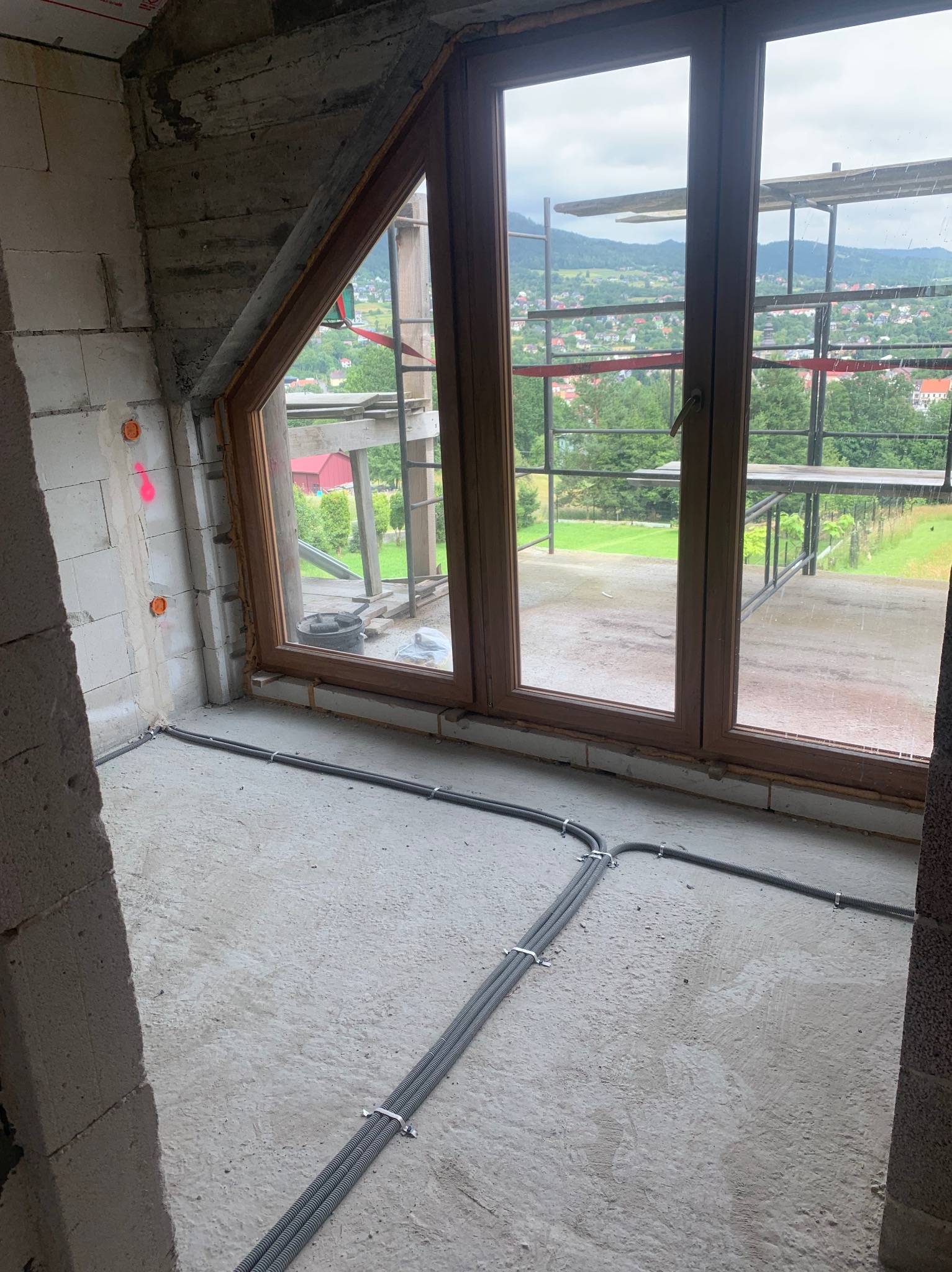 Surowe wnętrze w trakcie prac wykończeniowych, widoczne trzy kable w peszlach przytwierdzone do podłoża, okrągłe puszki instalacyjne w ścianie z betonu komórkowego oraz widok na góry przez okno.