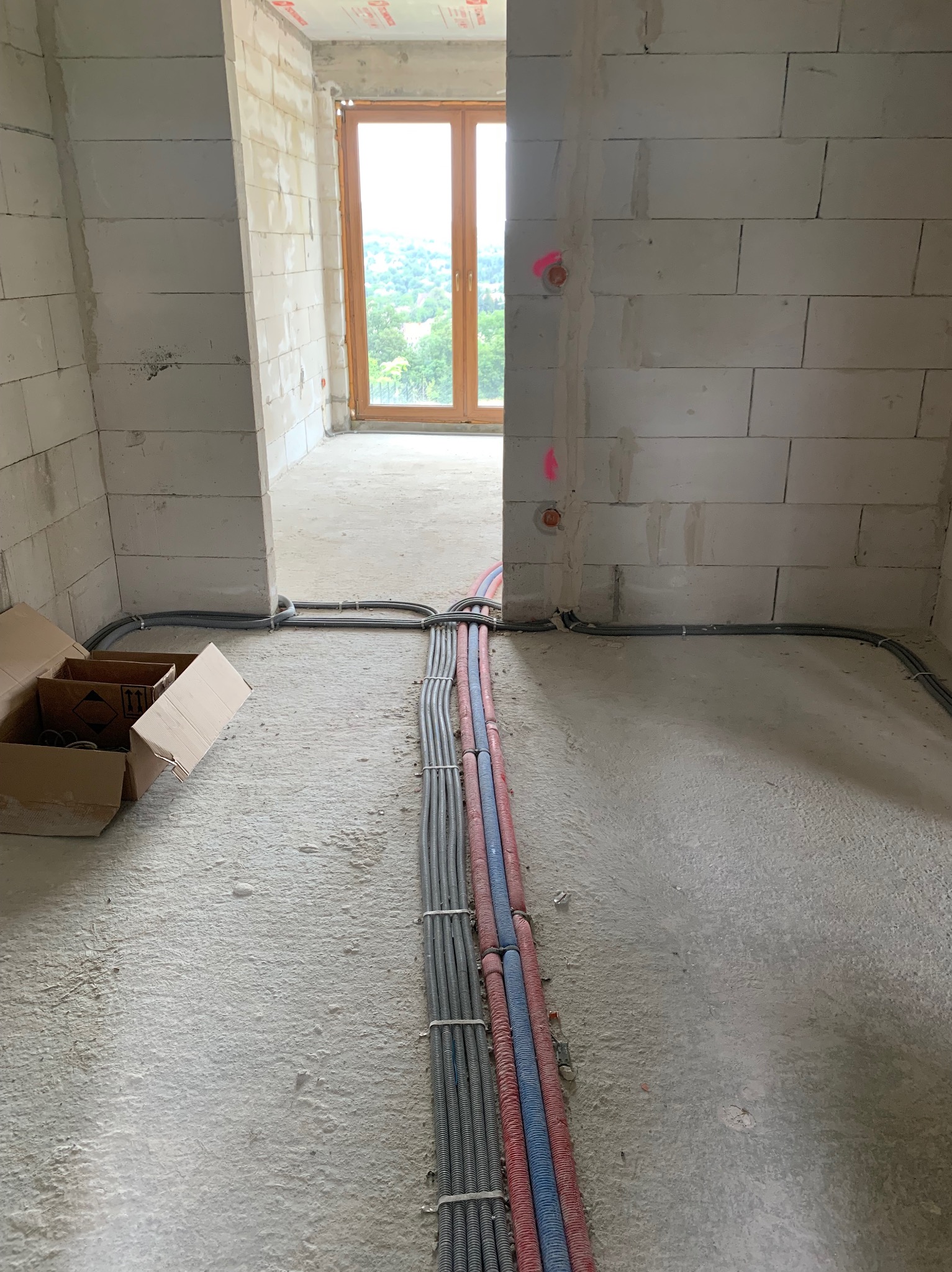 Wnętrze w stanie surowym z widocznymi przewodami elektrycznymi ułożonymi na podłodze i wyprowadzonymi na ścianach, oznaczone czerwonymi punktami. Część przewodów w peszlach w kolorach szarym...