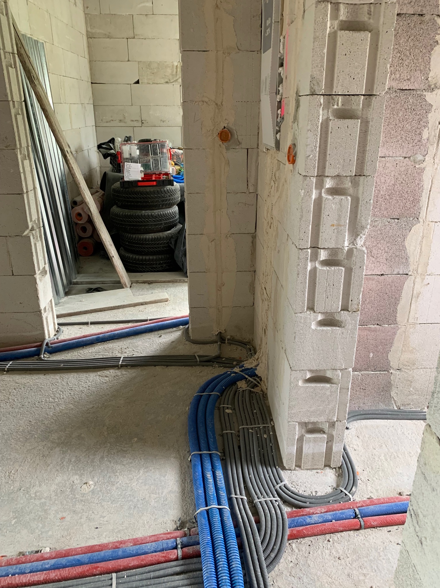 Wnętrze w trakcie budowy z widocznymi przewodami elektrycznymi w peszlach w kolorach niebieskim, szarym i czerwonym, ułożonymi na surowej podłodze wzdłuż ściany z betonu komórkowego. W tle widoczne...