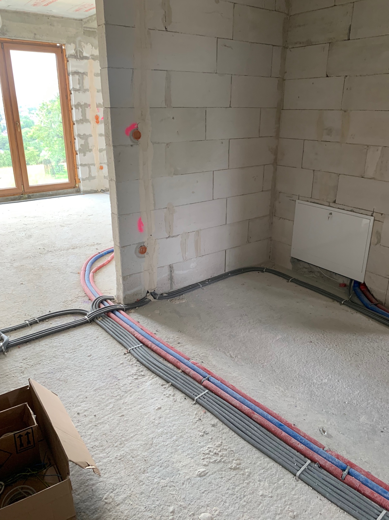 Wnętrze w stanie surowym z widocznymi przewodami elektrycznymi w peszlach ułożonymi na podłodze, prowadzącymi do ściany z widocznymi puszkami instalacyjnymi oznaczonymi różowym kolorem.
