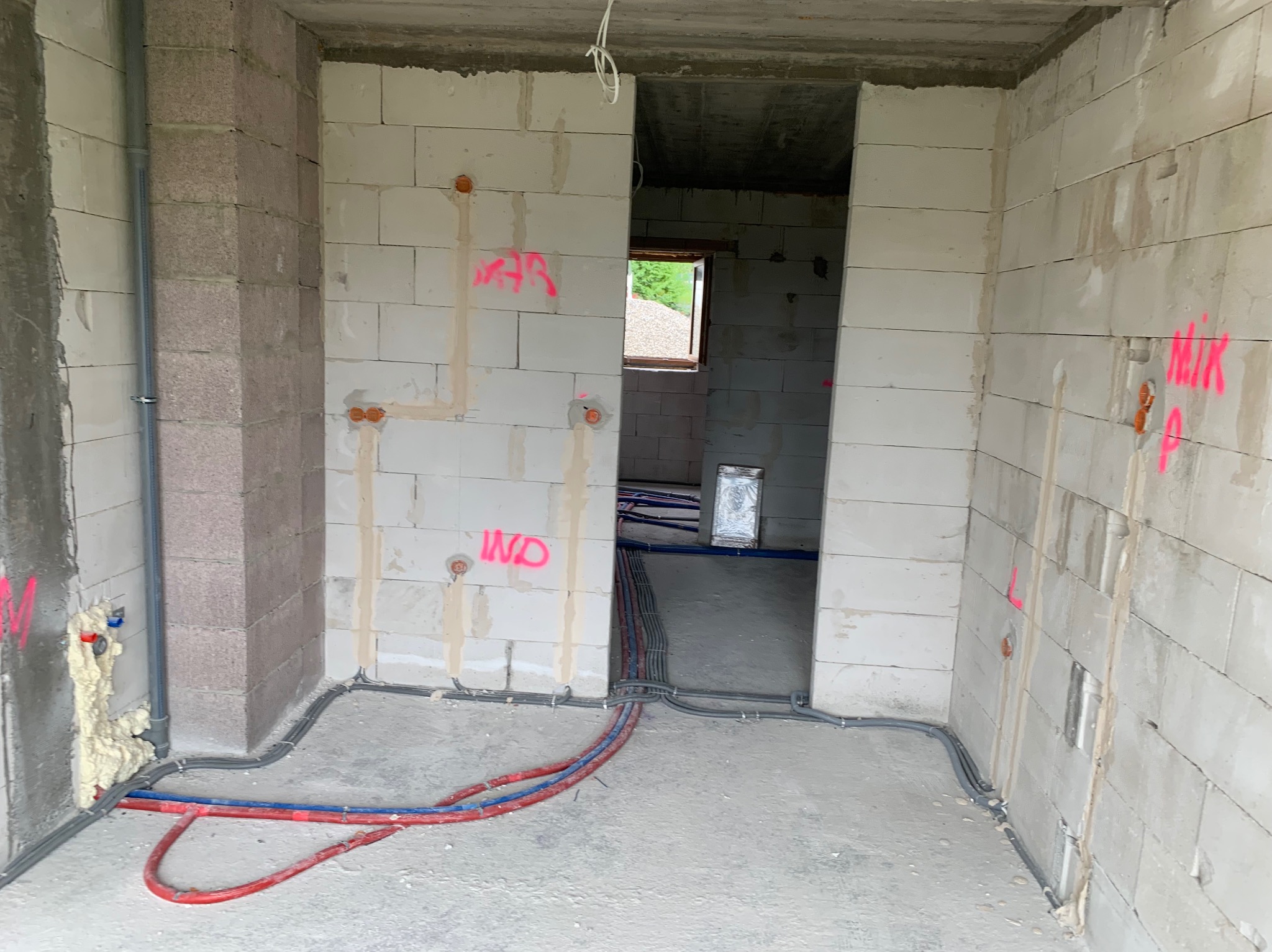 Wnętrze w stanie surowym z instalacją elektryczną i hydrauliczną w trakcie montażu, widoczne kable elektryczne i rury wodne prowadzone po podłodze i ścianach z betonu komórkowego, oznaczenia różową...