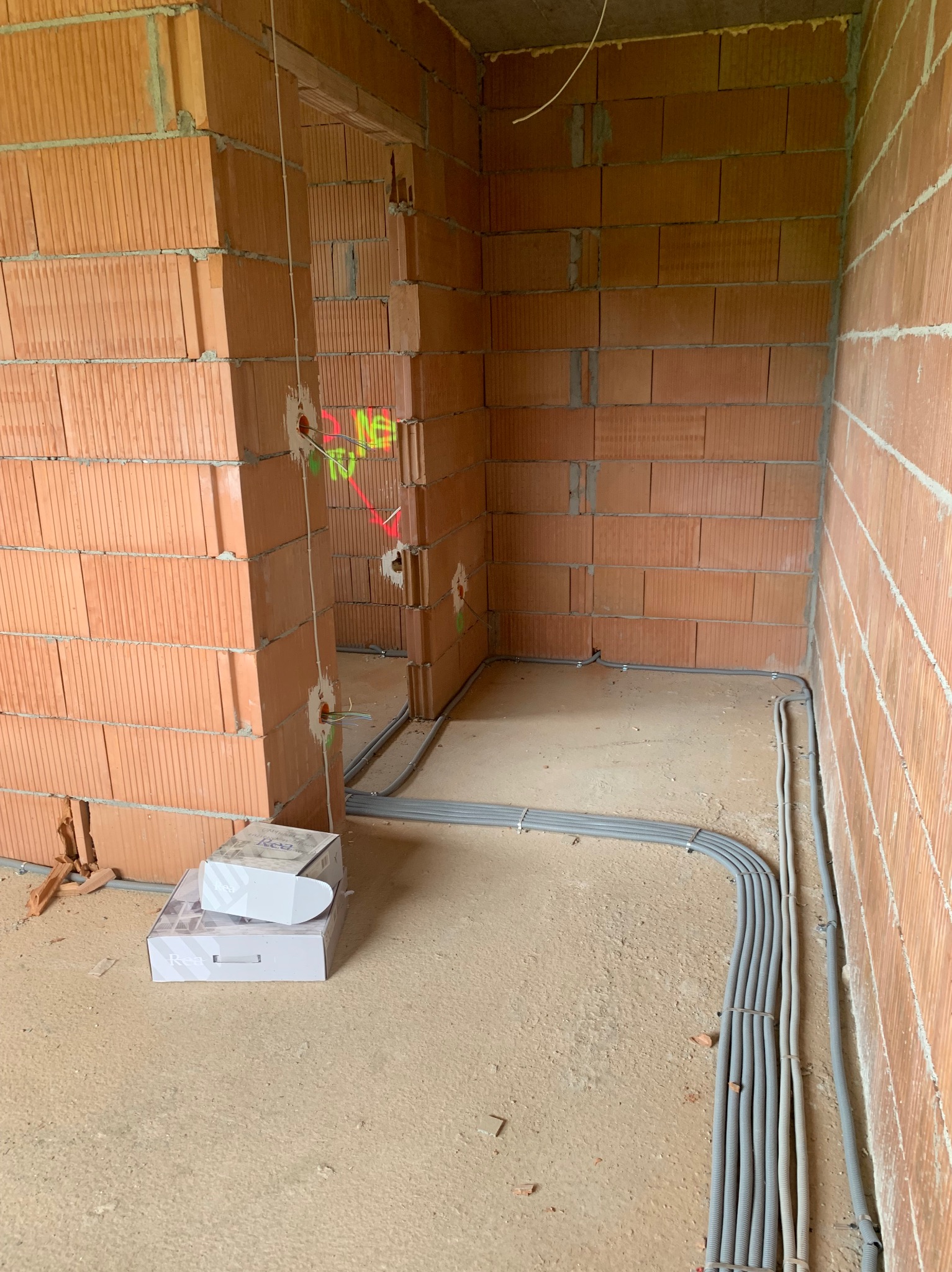Świeżo położone szare rury karbowane z przewodami elektrycznymi prowadzone po podłodze i ścianach z czerwonej cegły dziurawki w surowym stanie budynku, widoczne puszki instalacyjne z wystającymi...