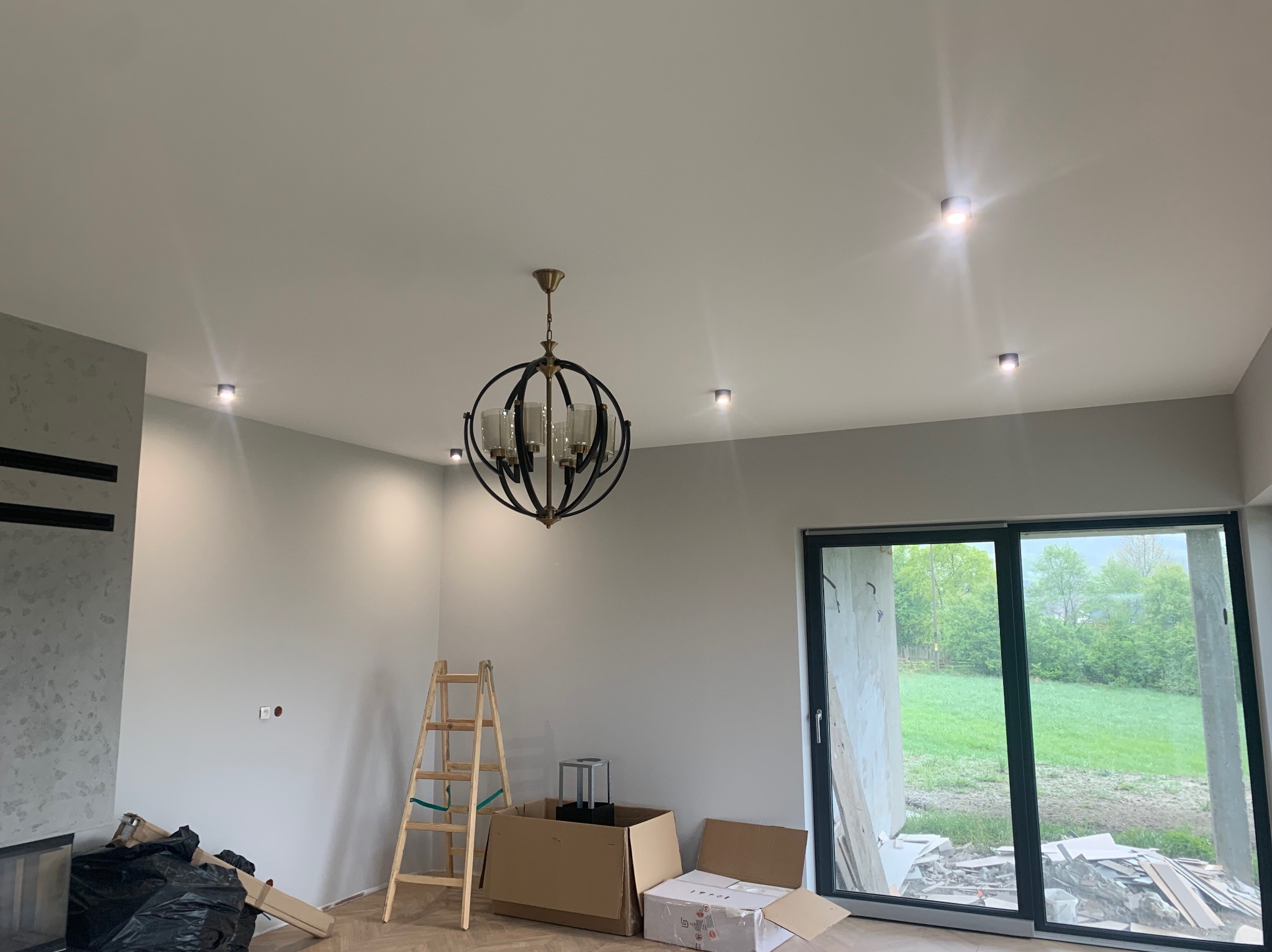 Wnętrze pokoju w trakcie wykańczania: zamontowany żyrandol w stylu loft, cztery punktowe lampy sufitowe, drabina oparta o ścianę, kartony na podłodze, widok na zewnątrz przez duże okno przesuwne.