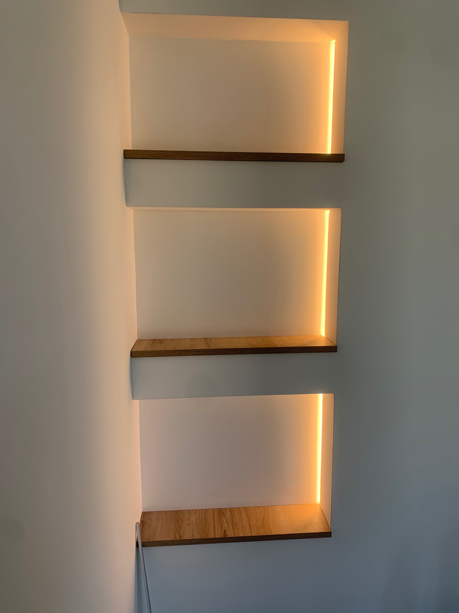 Wnęki ścienne z drewnianymi półkami i pionowym oświetleniem LED, ciepła barwa światła, minimalistyczny design.