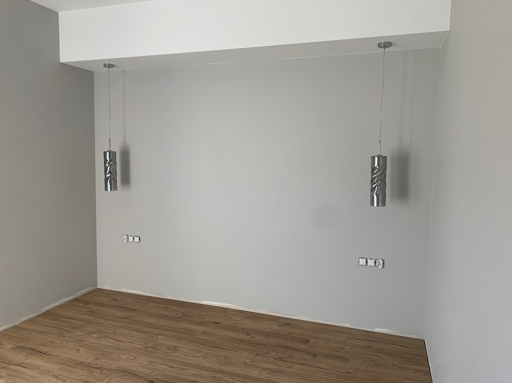 Dwa nowoczesne, srebrne, wiszące cylindryczne klosze lamp z dekoracyjnym wzorem, zamontowane na cienkich linkach w szarym pokoju z panelami podłogowymi, widoczne gniazdka elektryczne bez osprzętu.