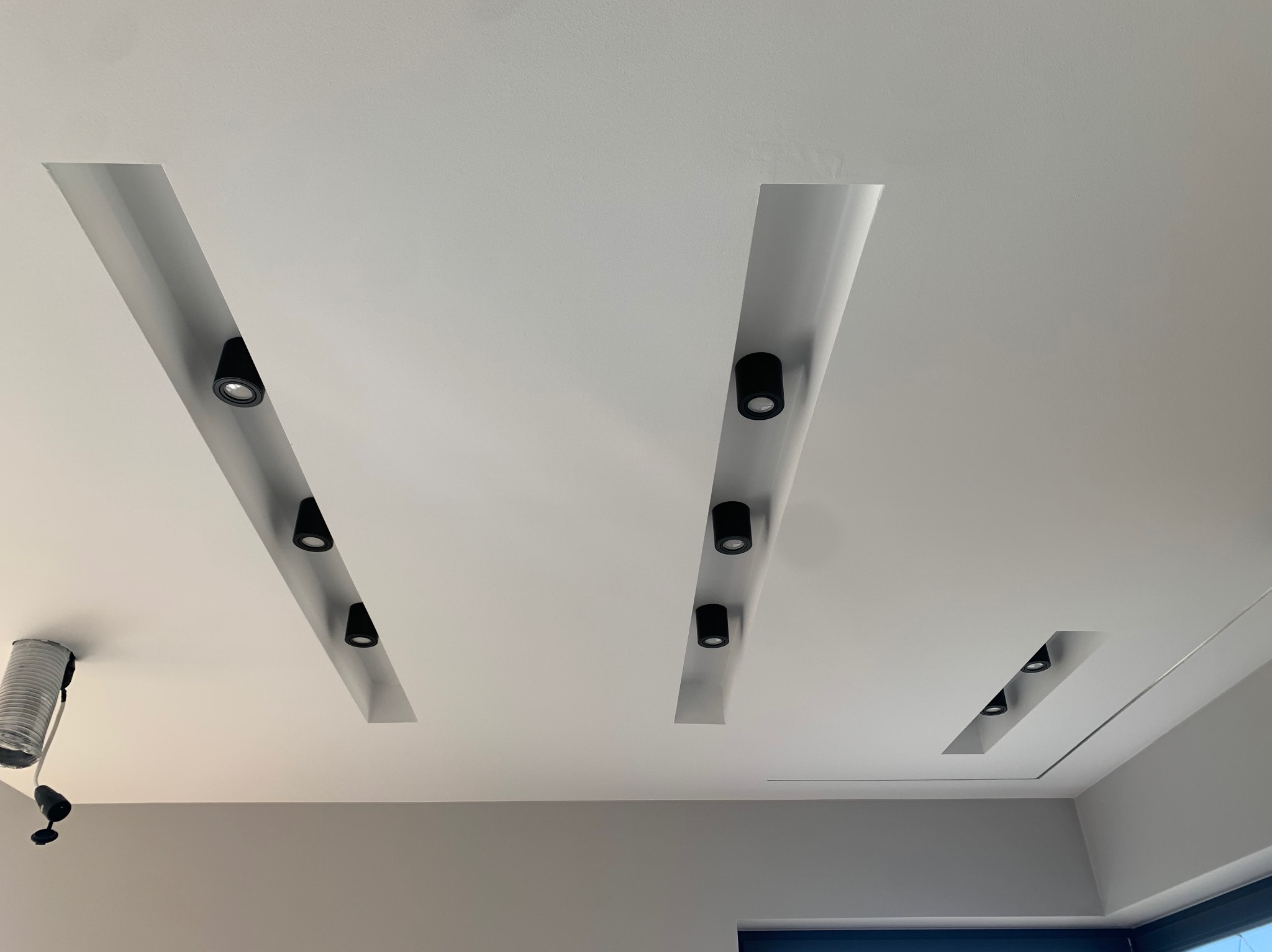Biały sufit z trzema wnękami oświetleniowymi, w każdej zamontowane czarne, punktowe reflektory LED. Po lewej stronie widoczna elastyczna rura wentylacyjna.